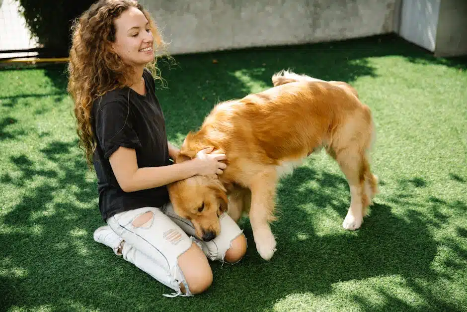 A smiling young woman with curly hair sitting on artificial grass and petting a golden retriever dog on a sunny day.